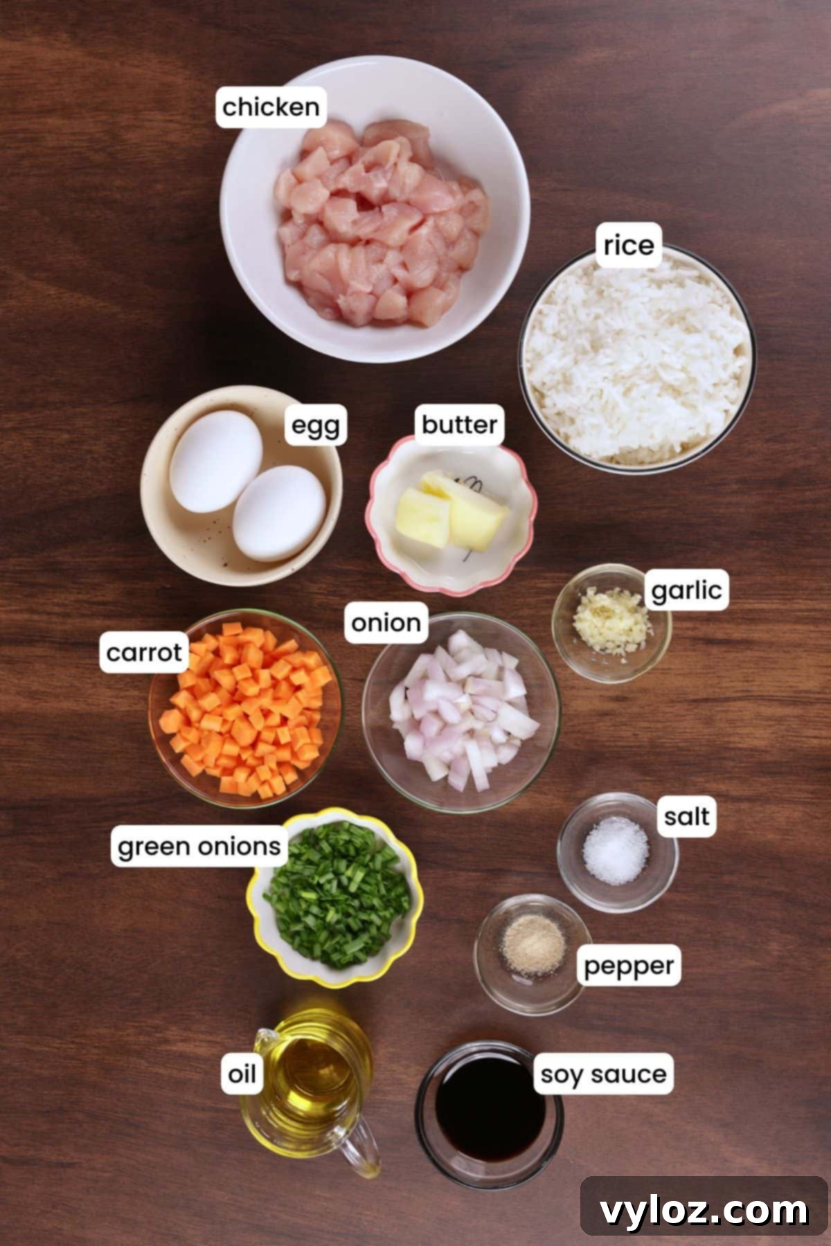 Ingredients for hibachi chicken fried rice laid out on a wooden background including rice, chicken, eggs, garlic, soy sauce, carrot, onion, and butter.