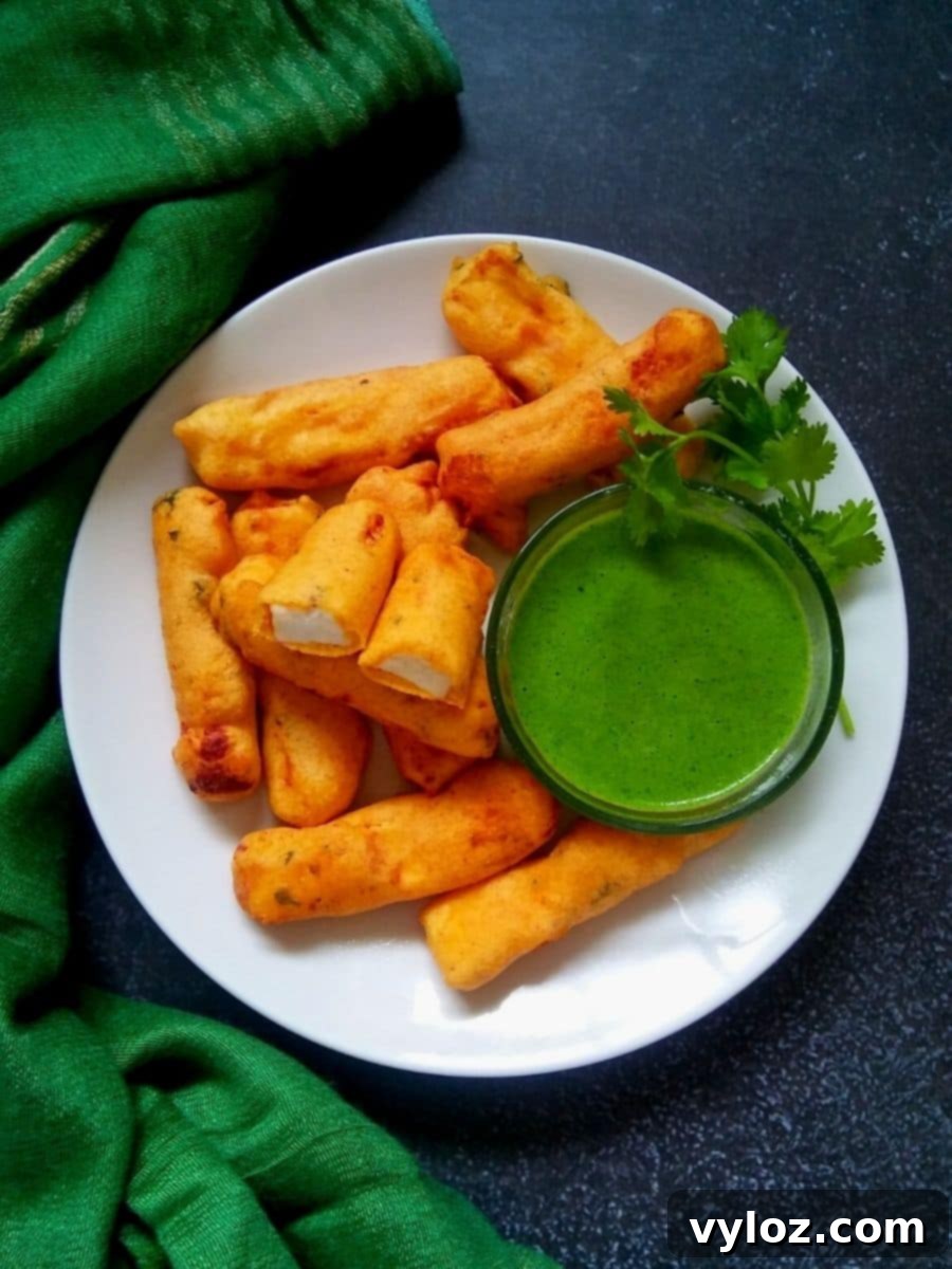 Crispy Paneer Pakora Made Easy 2 Crispy Paneer Pakora fritters served on a white plate with a side of vibrant green chutney in a bowl