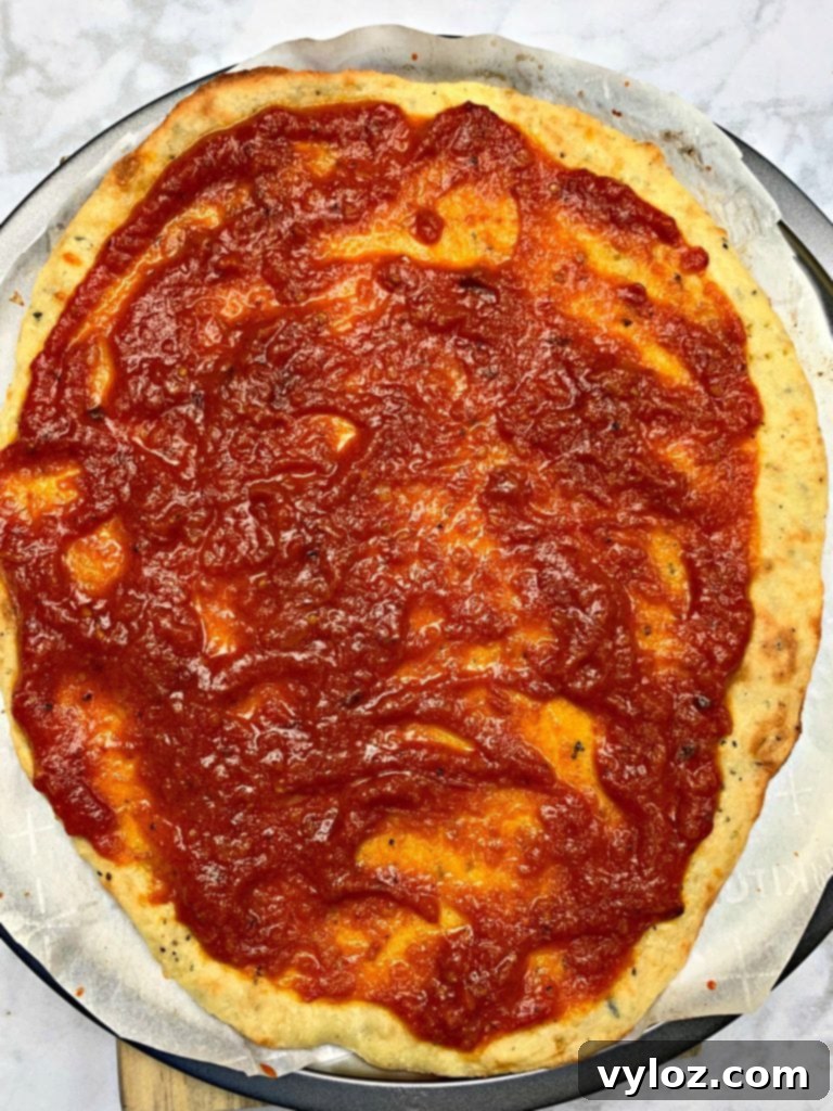 Pre-baked keto pepperoni pizza crust on a pizza pan with a thin, even layer of marinara sauce spread over it.