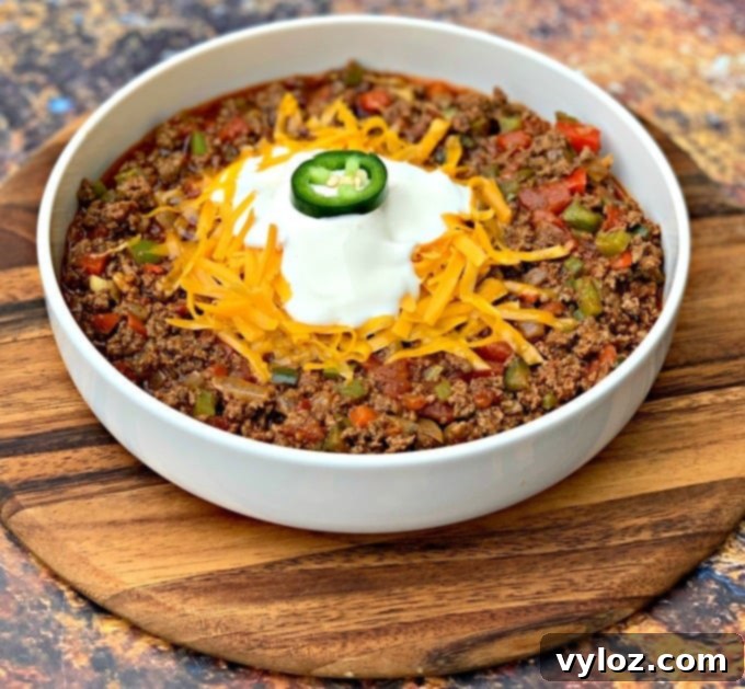 A generous serving of keto beef chili, richly topped with shredded cheese and a dollop of sour cream, presented in a pristine white bowl.