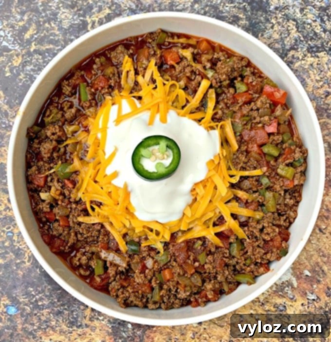 A close-up shot of keto beef chili served in a white bowl, generously garnished with melted shredded cheese.