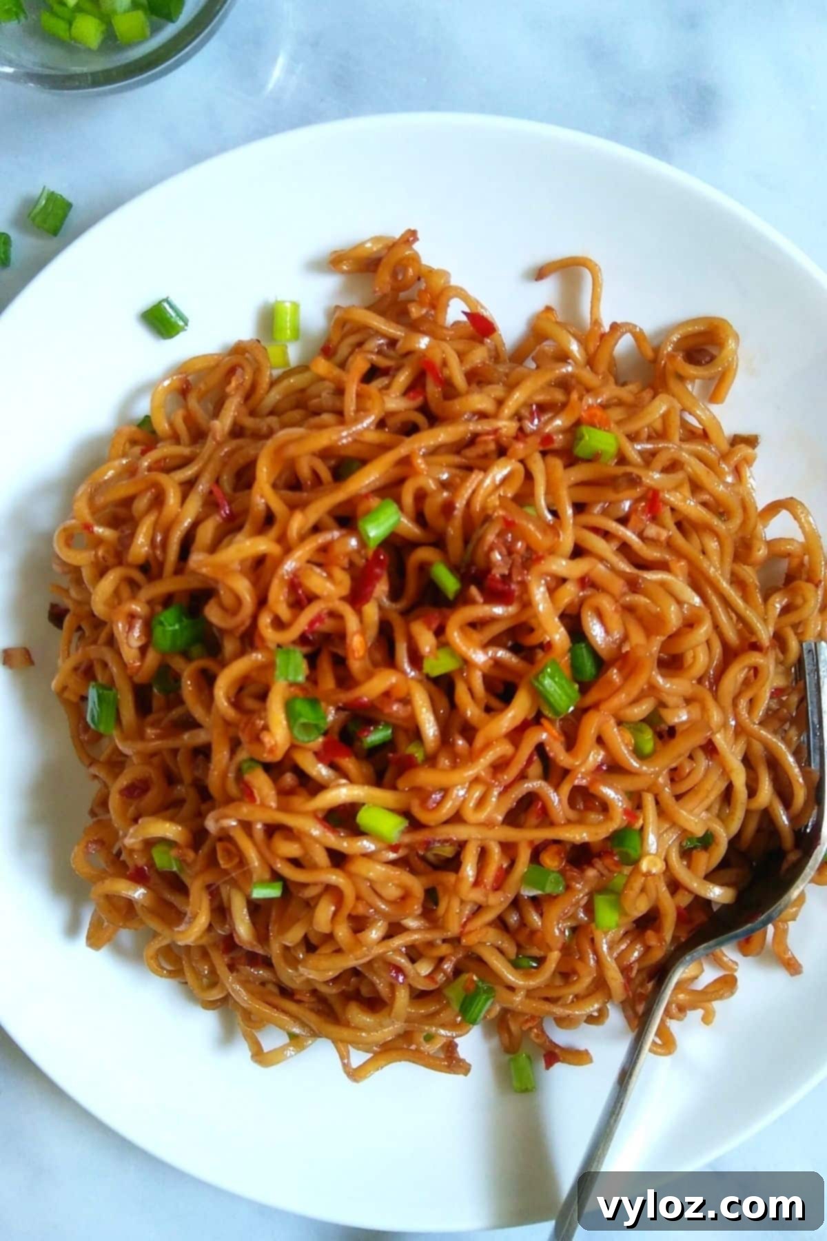 A vibrant bowl of spicy ramen noodles, garnished with fresh green onions and chili flakes, served on a pristine white plate, ready to be enjoyed.