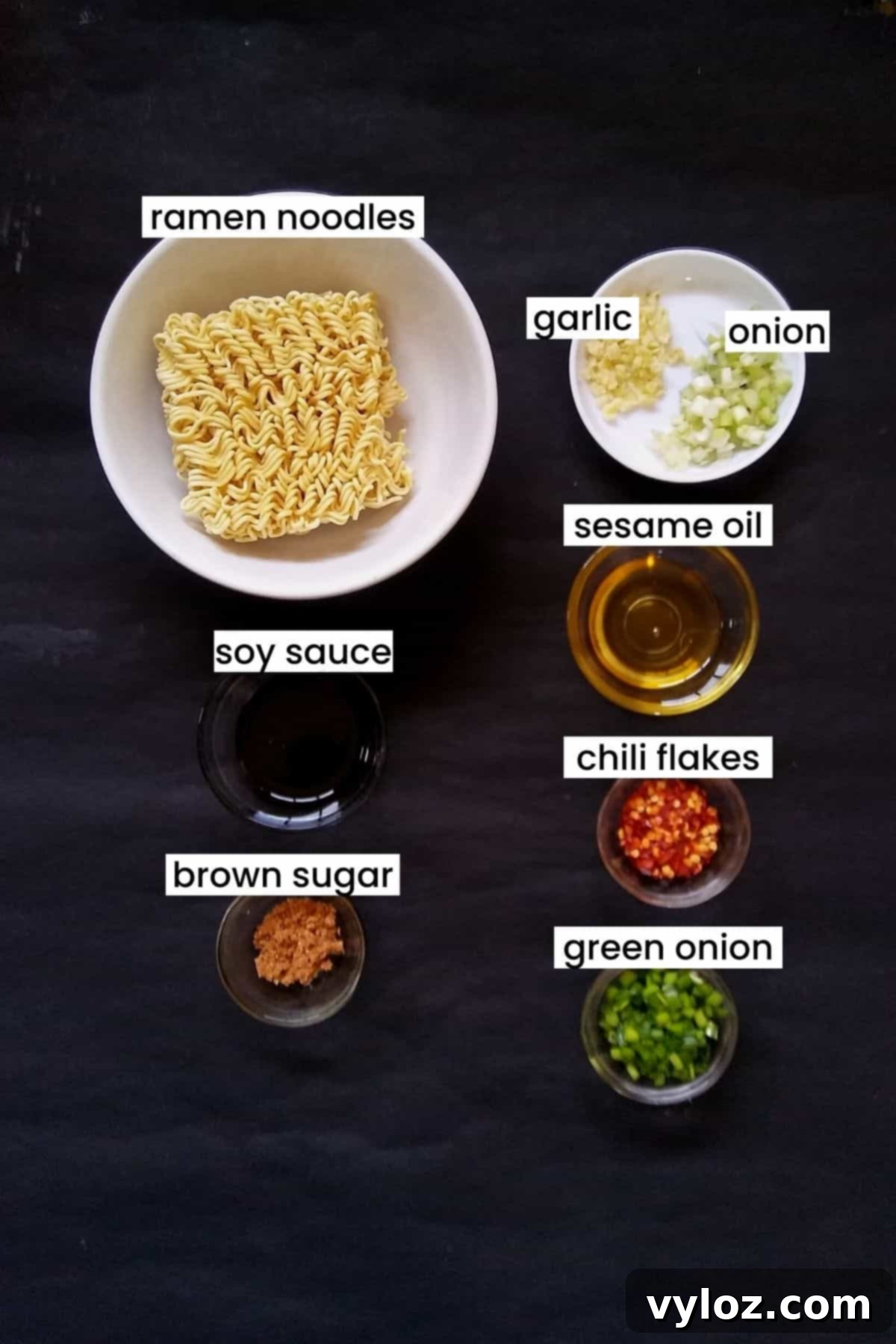 A beautifully arranged spread of ingredients for spicy ramen noodles, including instant ramen packets, fresh garlic, green onions, sesame oil, soy sauce, brown sugar, and red chili flakes, all set against a sleek black background.