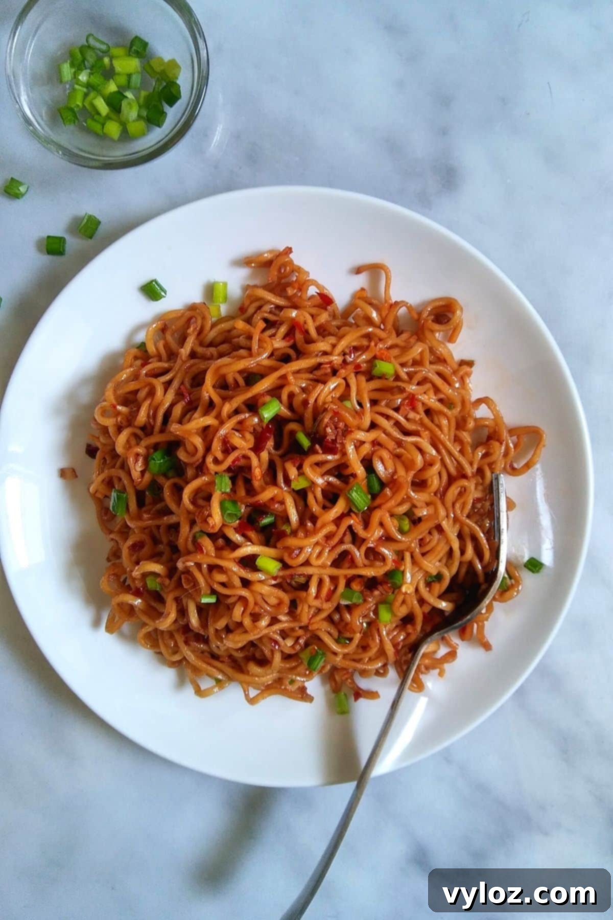 A close-up shot of a beautifully presented bowl of spicy ramen noodles, garnished with vibrant green onions, served on a clean white plate, ready to be savored.
