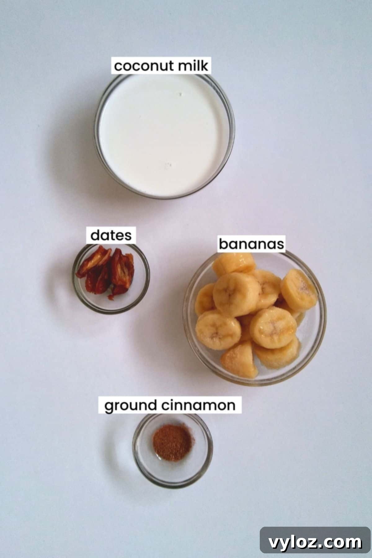 Ingredients for a healthy banana milkshake: coconut milk, pitted dates, sliced frozen bananas, and ground cinnamon on a wooden board.