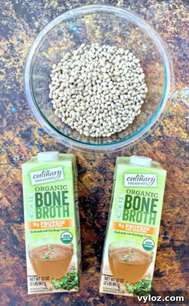 Raw navy beans in a glass bowl and bottles of chicken broth