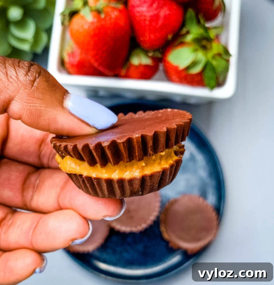 A hand holding a perfectly formed sugar-free peanut butter cup, with a bowl of fresh strawberries blurred in the background.