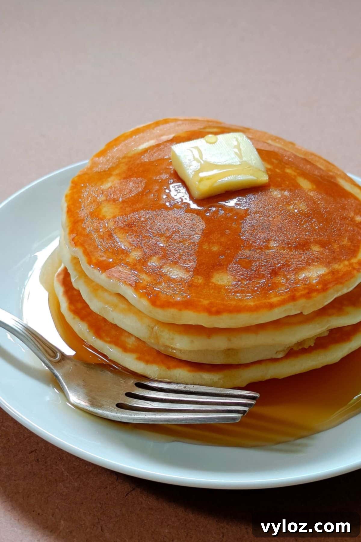 Image showing golden fluffy pancakes stacked on a white plate, drizzled with maple syrup and a pat of butter.