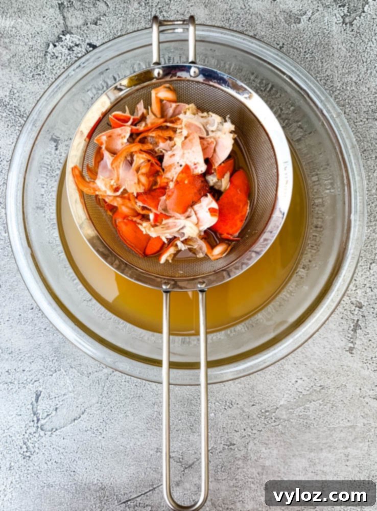 A close-up shot of freshly shelled lobster meat resting in a fine-mesh strainer over a bowl, with clear, golden lobster stock in the background, symbolizing the process of separating flavorful broth from the succulent meat.