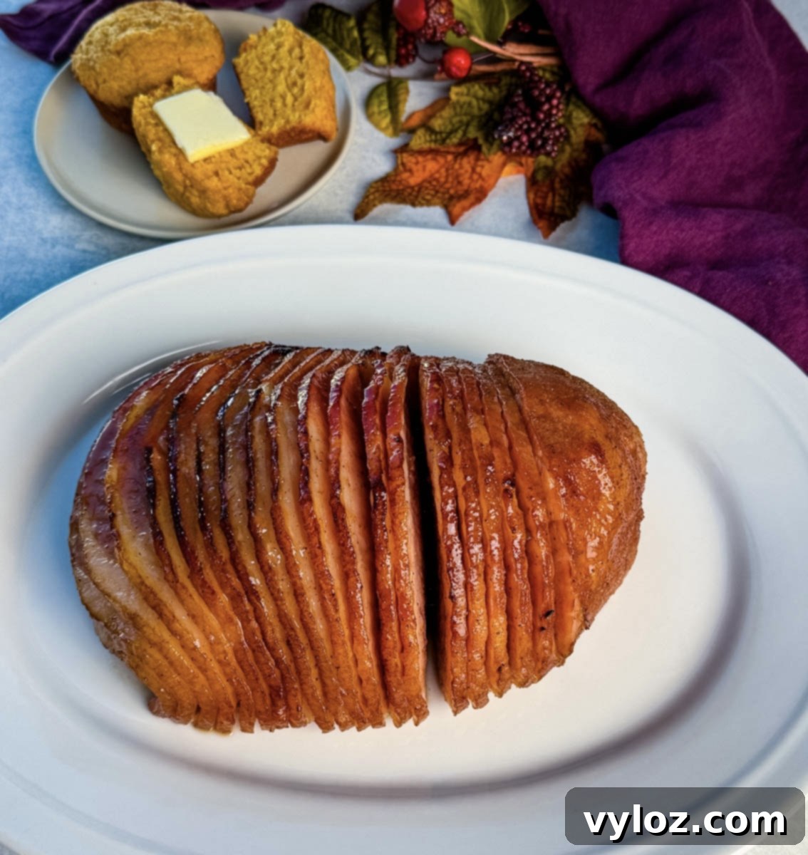 A full view of a beautifully prepared spiral honey-glazed ham on a pristine white serving platter, complemented by freshly baked cornbread muffins, a bowl of butter, and a warm, fall-themed background decor, inviting guests to a festive meal.