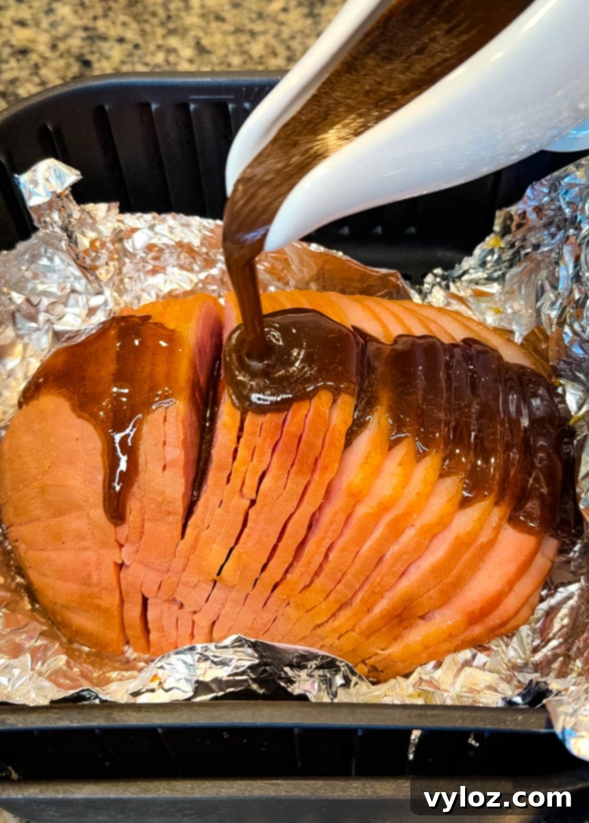 Thick, rich brown glaze is generously poured over a pre-sliced ham, which is nestled in foil inside an air fryer basket, ensuring every crevice is coated for optimal caramelization.