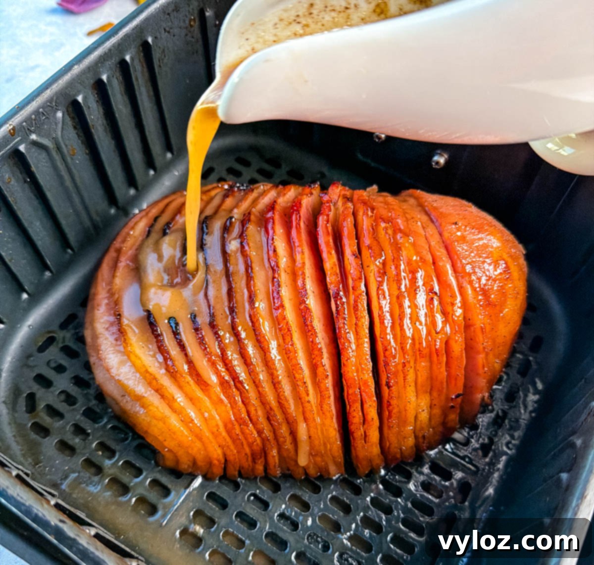 A pristine white pitcher pours a generous stream of golden honey glaze over a spiral ham, carefully placed inside an air fryer basket, ensuring a uniform and glossy coating.