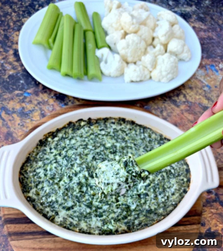 Warm Baked Spinach and Cheese Dip 8 Person holding a celery stick dipped in keto low carb spinach dip, with other vegetables on a white plate