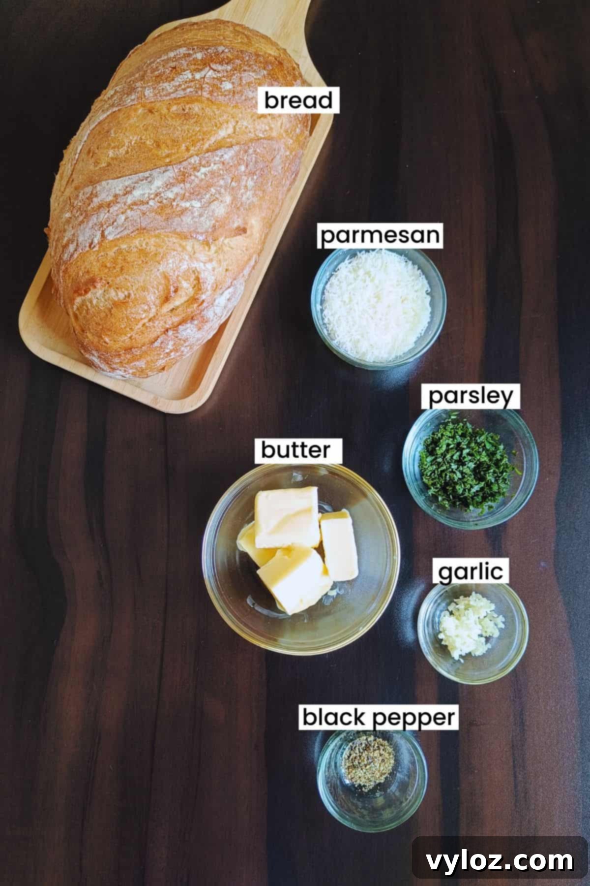 Garlic bread ingredients laid out on a rustic wooden surface, including a loaf of bread, butter, garlic cloves, parsley, and parmesan cheese.
