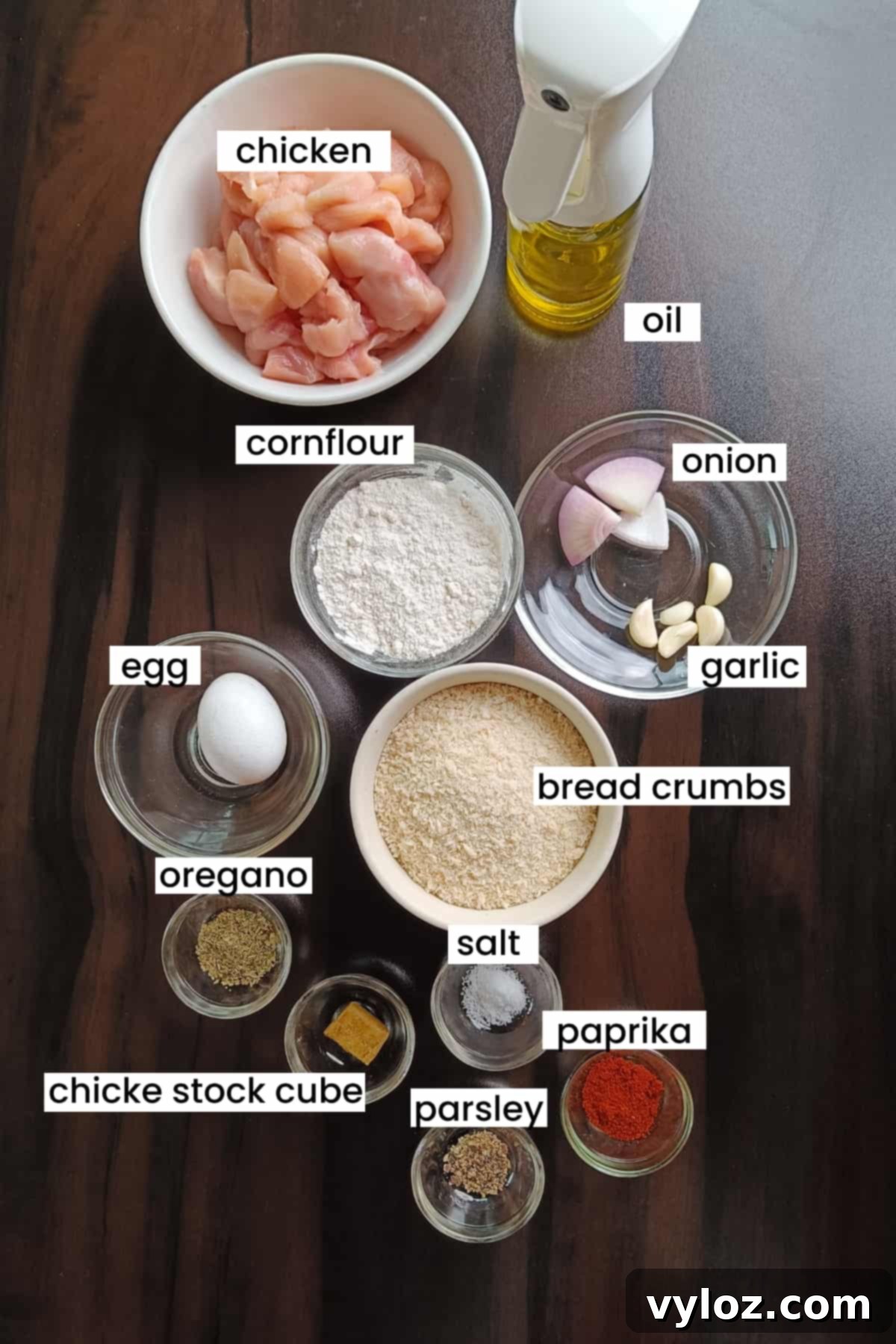 Image showing air fryer chicken nuggets ingredients like chicken breast, onion, garlic, spices, egg, cornflour, breadcrumbs, and oil placed on a wooden background.