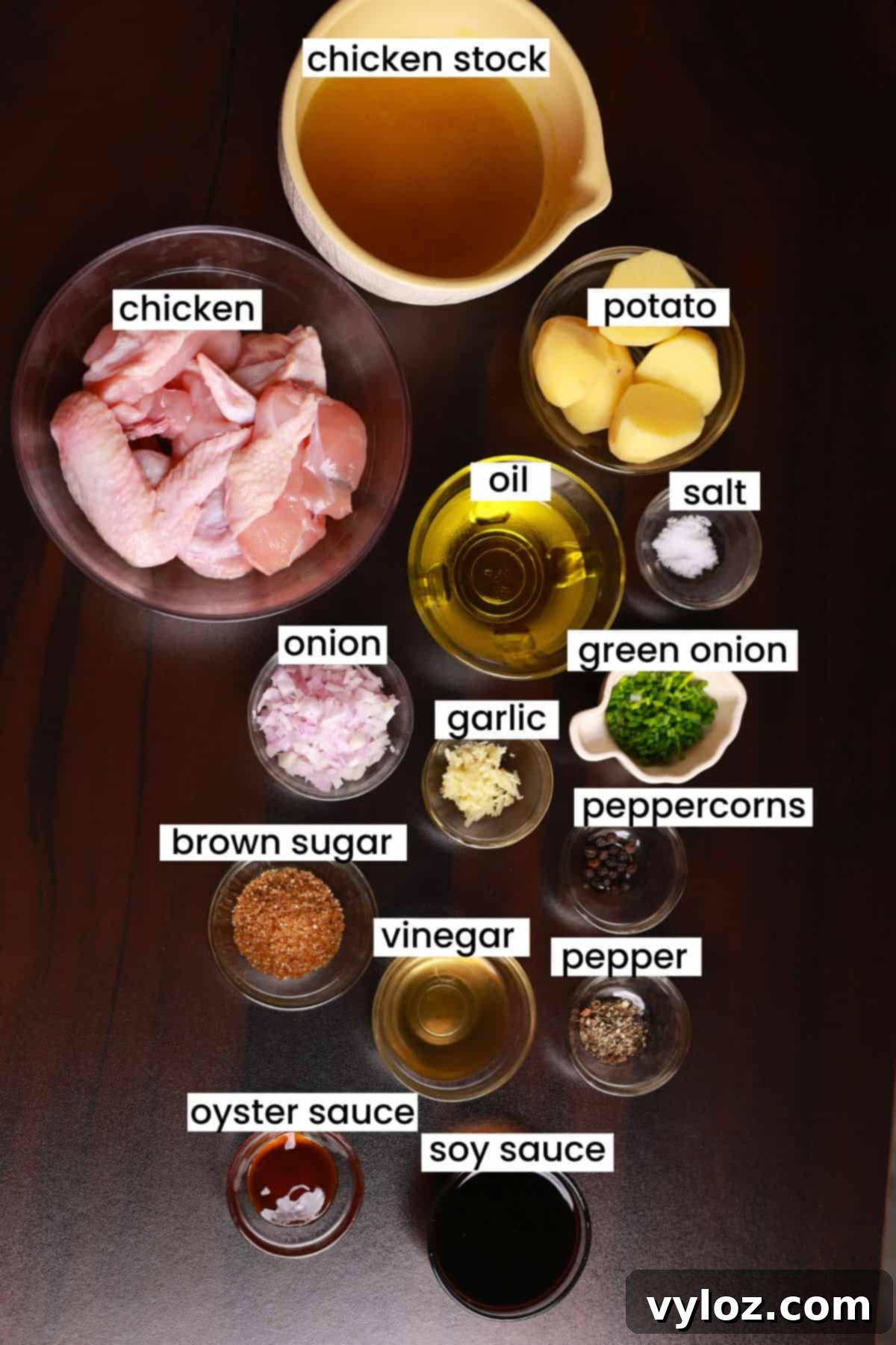 A collection of fresh ingredients for Filipino chicken adobo with potatoes, including chicken, potatoes, soy sauce, vinegar, garlic, and bay leaves, neatly arranged on a brown wooden background.