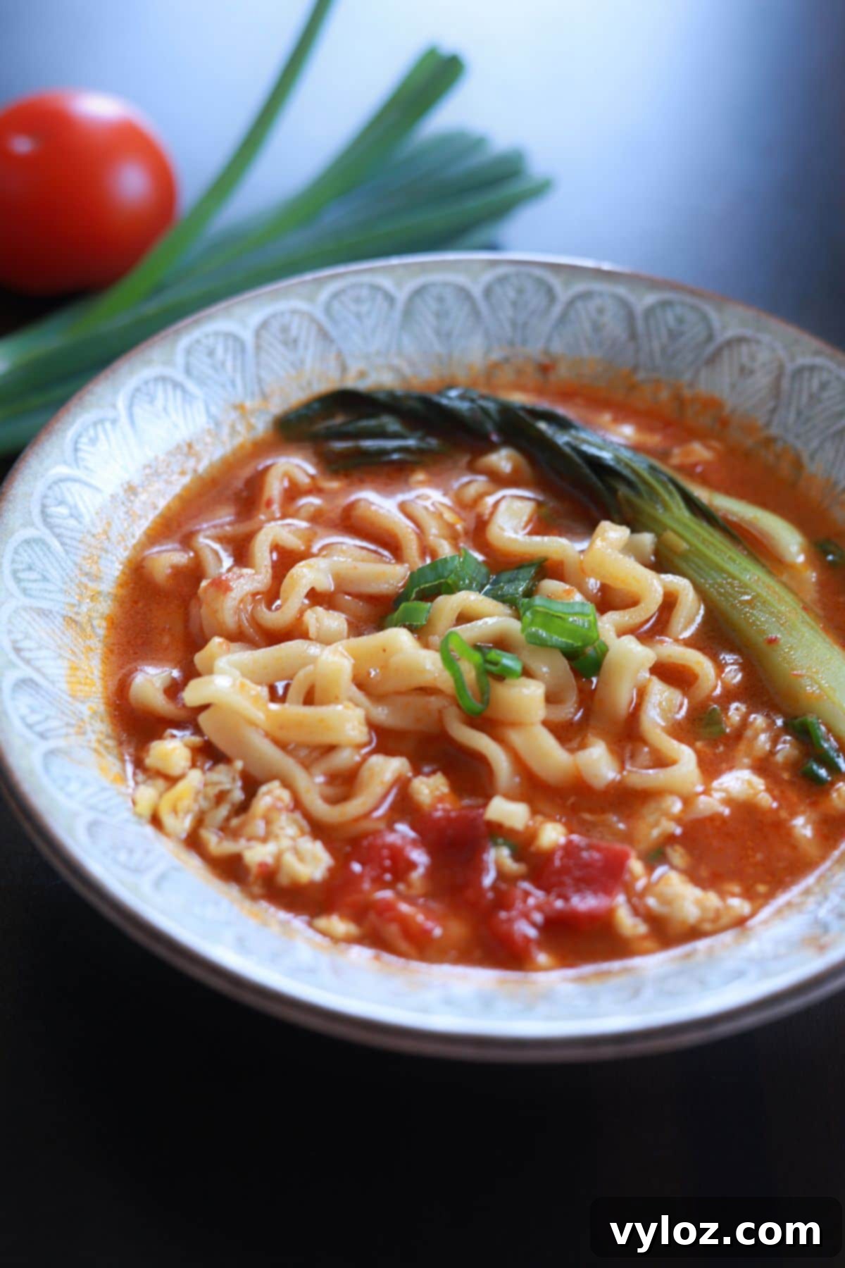 A vibrant bowl of 10-minute tomato ramen soup, showcasing a rich red broth, perfectly cooked instant noodles, fluffy scrambled egg, fresh baby bok choy, and a sprinkle of green onions for garnish.