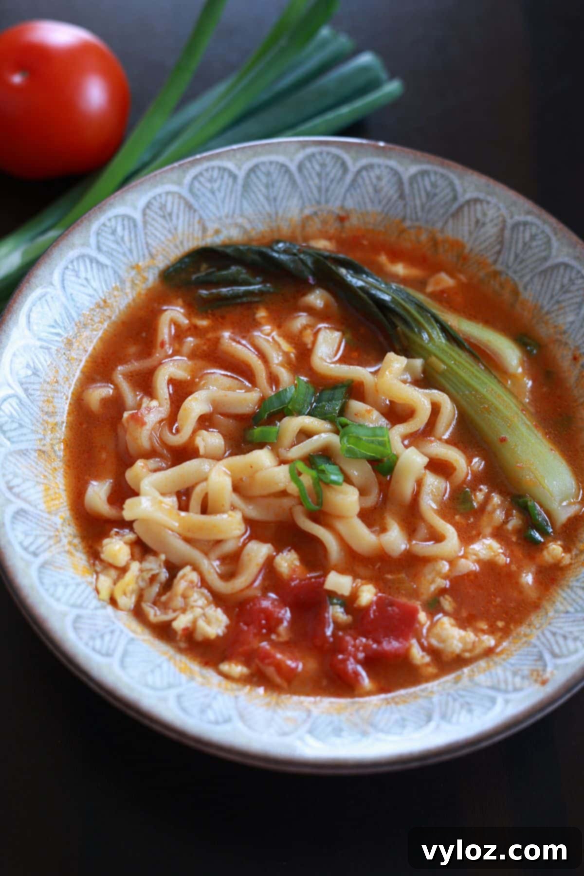A perfectly styled close-up of a bowl of 10-minute tomato ramen, featuring glossy instant noodles submerged in a vibrant red broth, topped generously with fluffy scrambled egg, tender baby bok choy, and bright green onions, ready to be enjoyed.