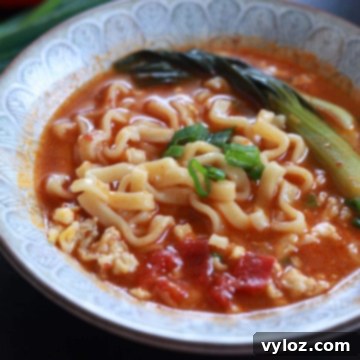 A bowl of tomato ramen with a rich, red broth, topped with scrambled egg, baby bok choy, and green onions.