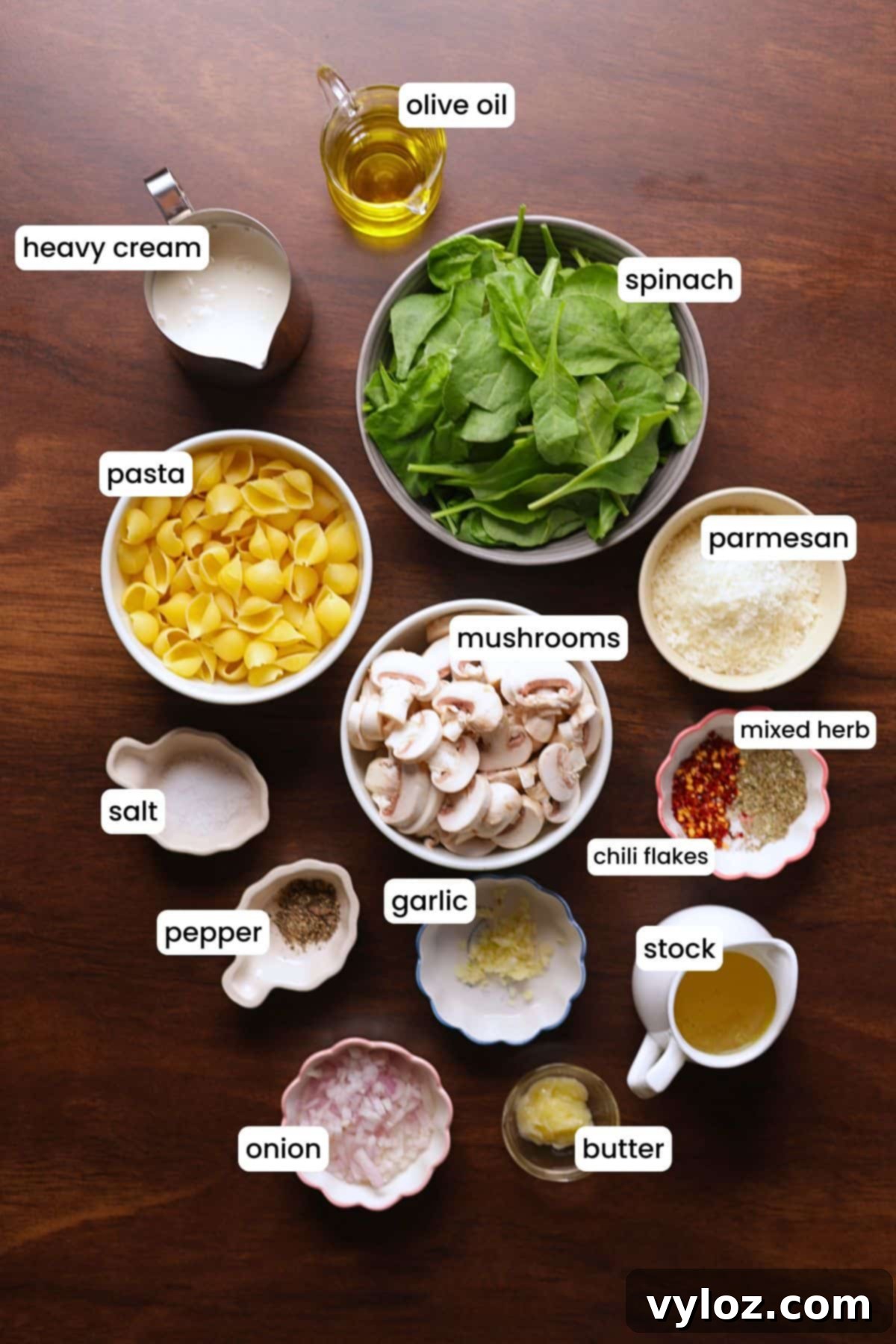 Velvety Mushroom and Spinach Pasta 3 All ingredients for creamy mushroom and spinach pasta arranged on a wooden background, including mushrooms, spinach, pasta shells, garlic, onion, olive oil, butter, chili flakes, Italian seasoning, stock, cream, and Parmesan cheese.