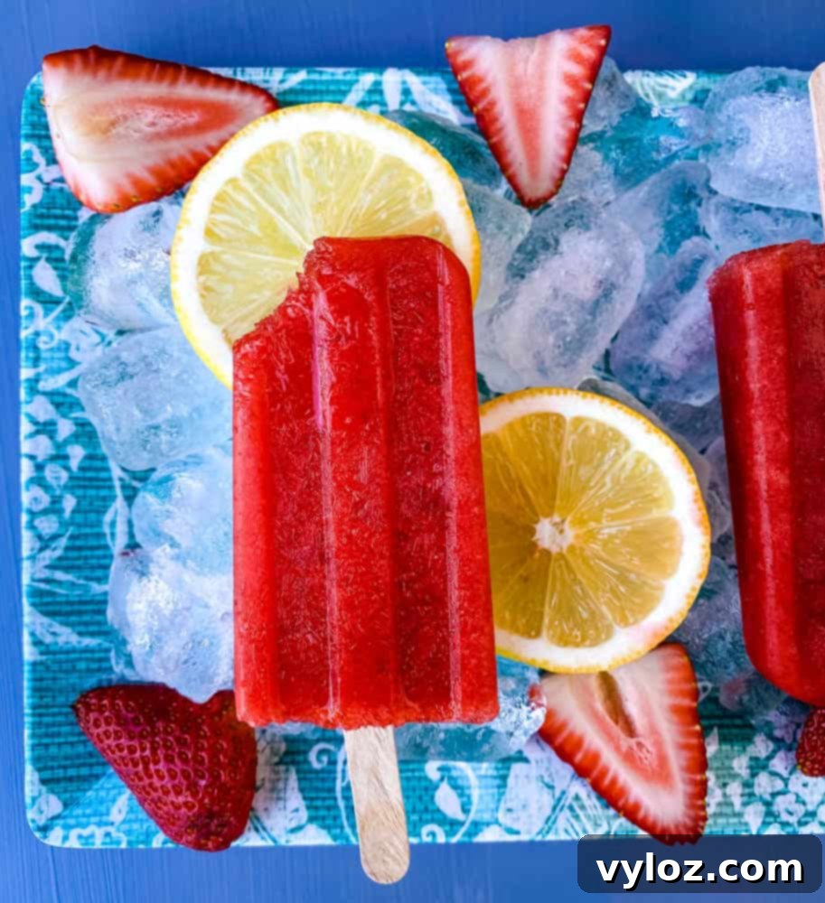 sugar free strawberry lemon popsicle with a bite removed on a flat surface with ice, fresh lemons and strawberries 
