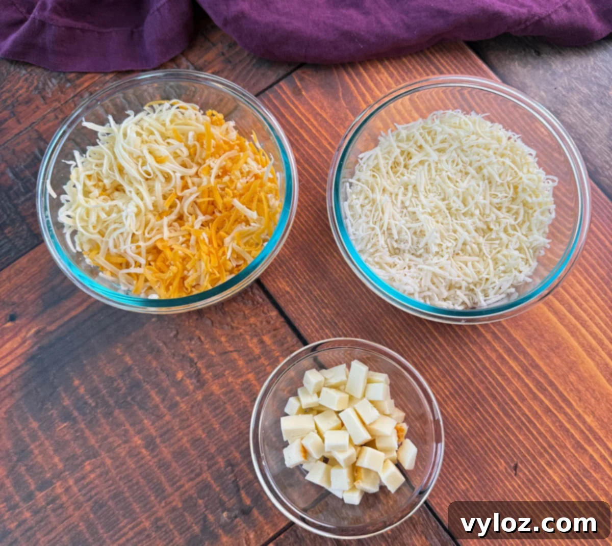 Soul-Satisfying Southern Baked Mac and Cheese 7 Overhead view of three glass bowls on a wooden table—one with shredded cheddar and mozzarella, one with shredded white cheese, and one small bowl with cubed cheese.