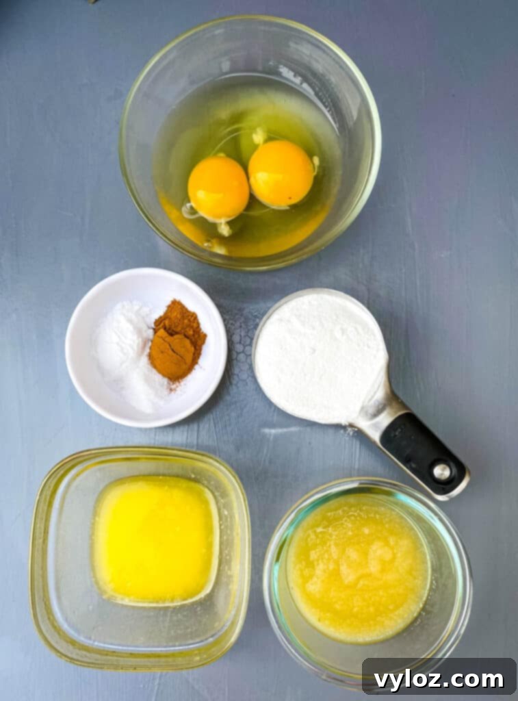 A collection of key ingredients for banana bread: raw eggs, cinnamon powder, a bowl of granular sweetener, melted butter, and unsweetened applesauce laid out on a flat surface