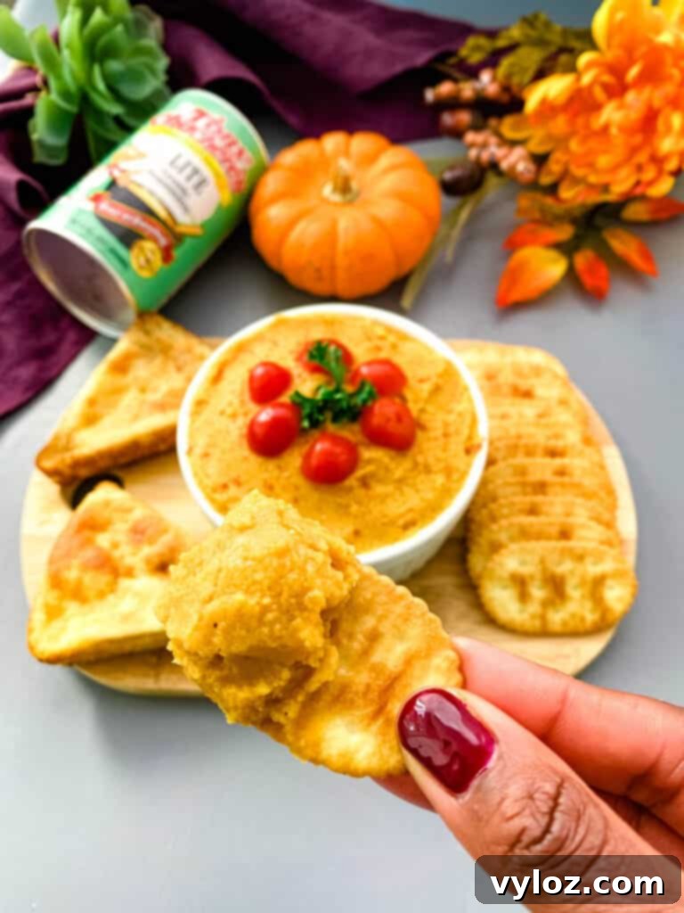 A hand holding a cracker generously topped with creamy pumpkin hummus, ready to be enjoyed.