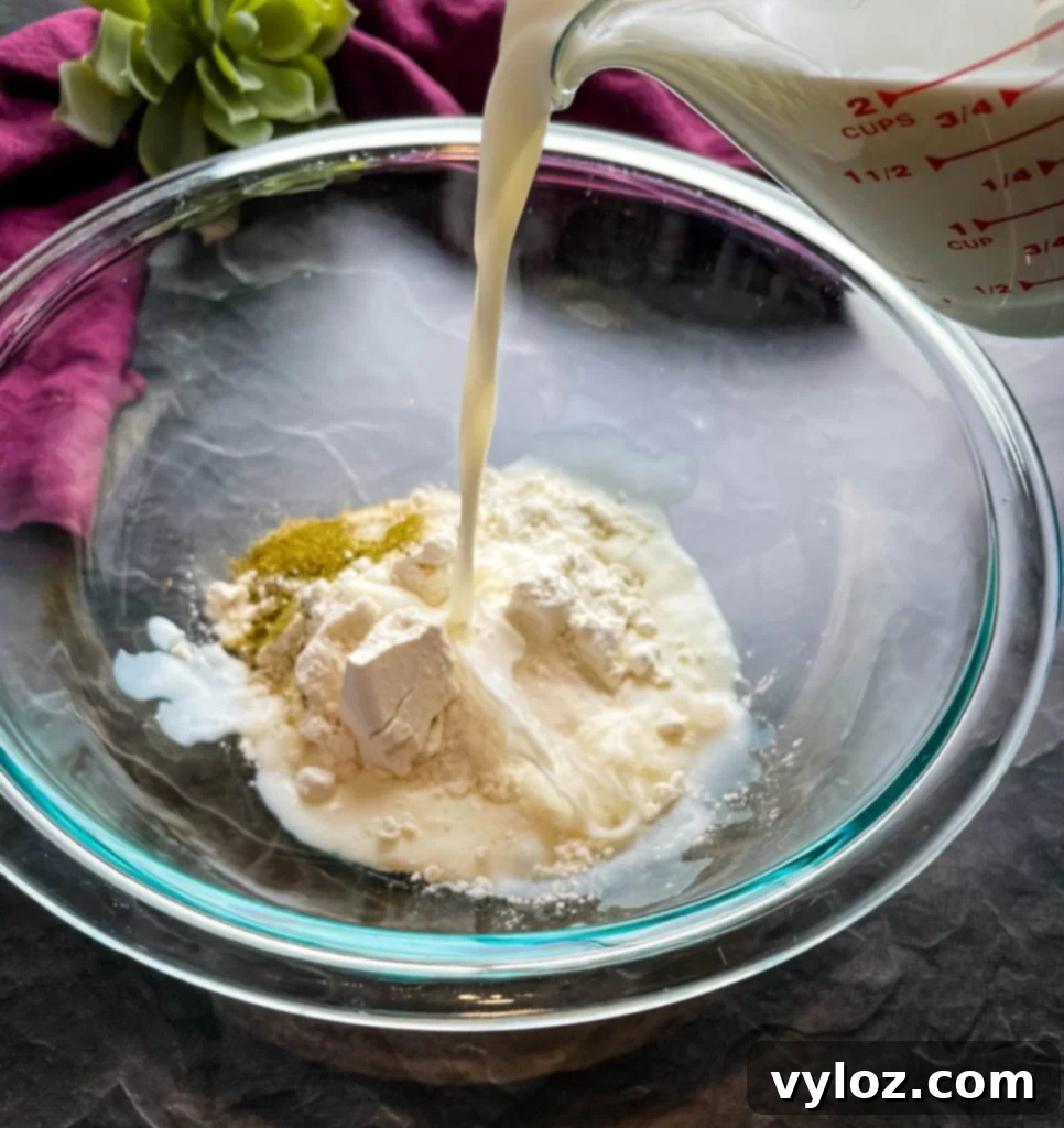 Milk being carefully poured into a clear glass bowl, joining flour and a variety of seasonings, ready to be whisked into a smooth mixture.