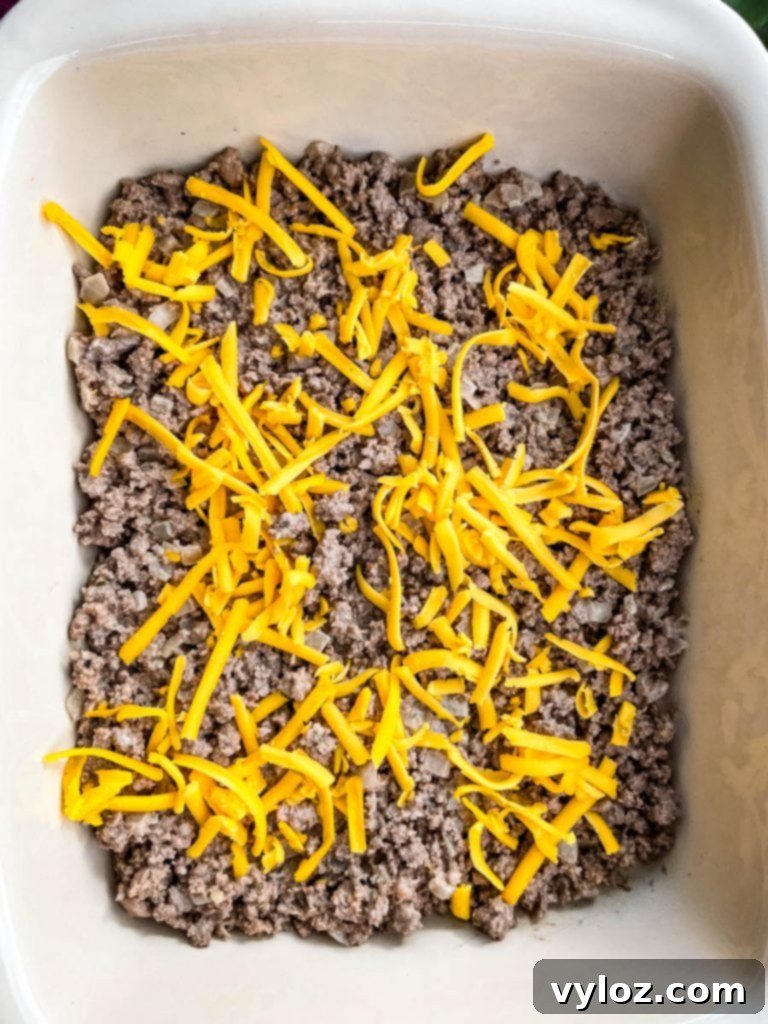 Big Mac Bake 8 The cooked ground beef layer in a casserole dish, topped with a generous sprinkle of shredded cheddar cheese, creating a delicious base for the sauce layer.