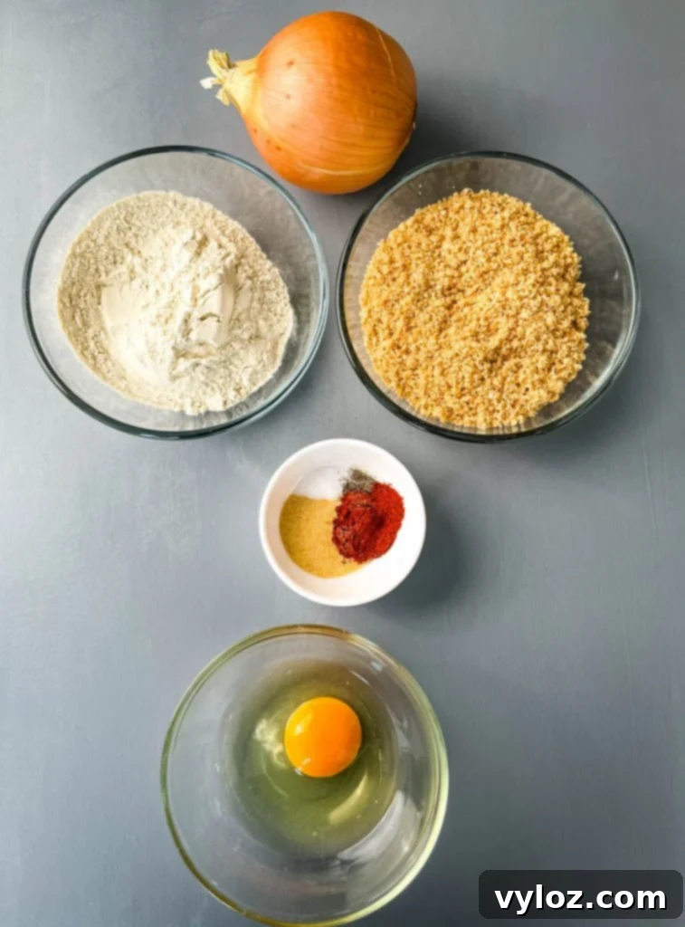 Crispy Keto Low-Carb Onion Rings 3 Arrangement of raw onion slices, finely crushed pork rinds, keto flour, a raw egg, and various seasonings laid out on a flat wooden surface, ready for preparing keto onion rings.