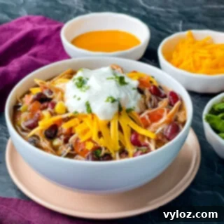 Tangy Buffalo Chicken Chili 12 A generously portioned white bowl of buffalo chicken chili, adorned with a sprinkle of shredded cheese and a dollop of cooling sour cream on top.