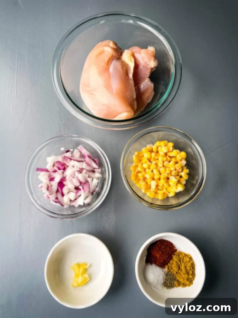 Tangy Buffalo Chicken Chili 4 Array of raw ingredients for buffalo chicken chili: fresh chicken breasts, finely chopped red onions, minced garlic, a bowl of frozen corn kernels, and a mix of chili seasoning, neatly arranged on a flat surface.