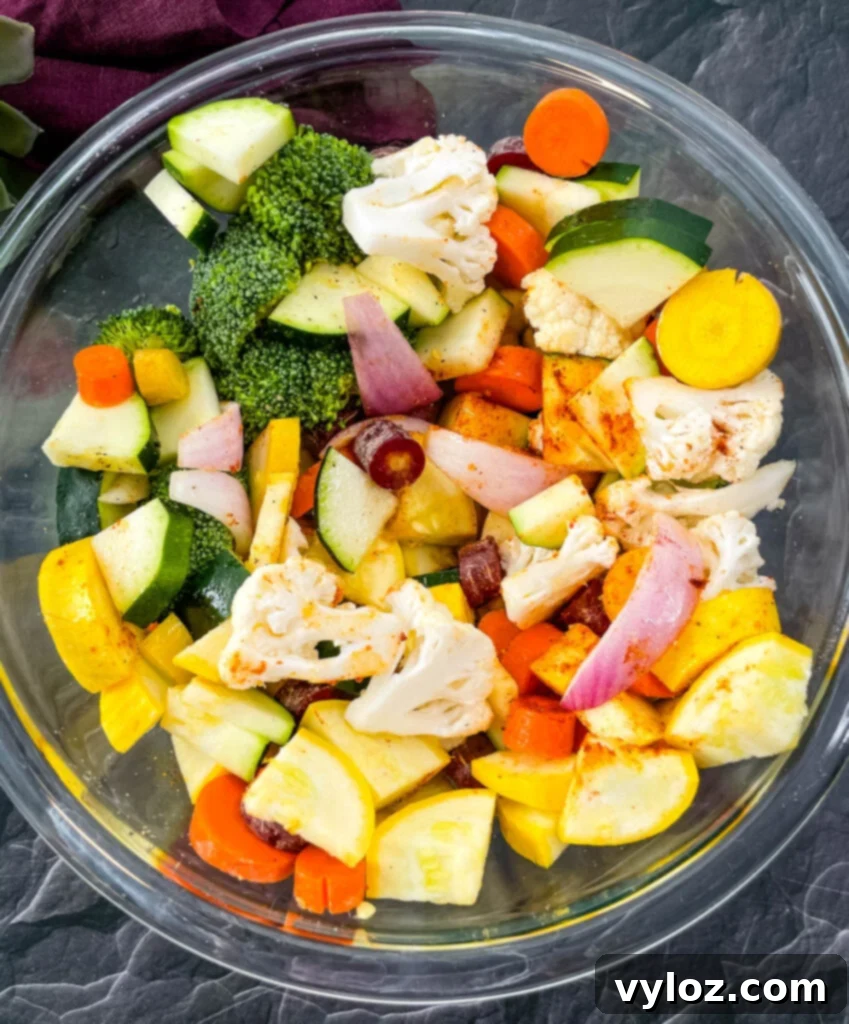 A glass bowl overflowing with squash, zucchini, onions, carrots, broccoli, and cauliflower, thoroughly coated in seasoning and marinade.