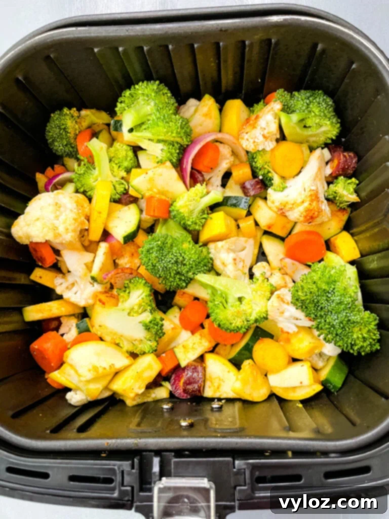A colorful assortment of squash, zucchini, onions, carrots, broccoli, and cauliflower arranged in a single layer in an air fryer basket, ready for cooking.