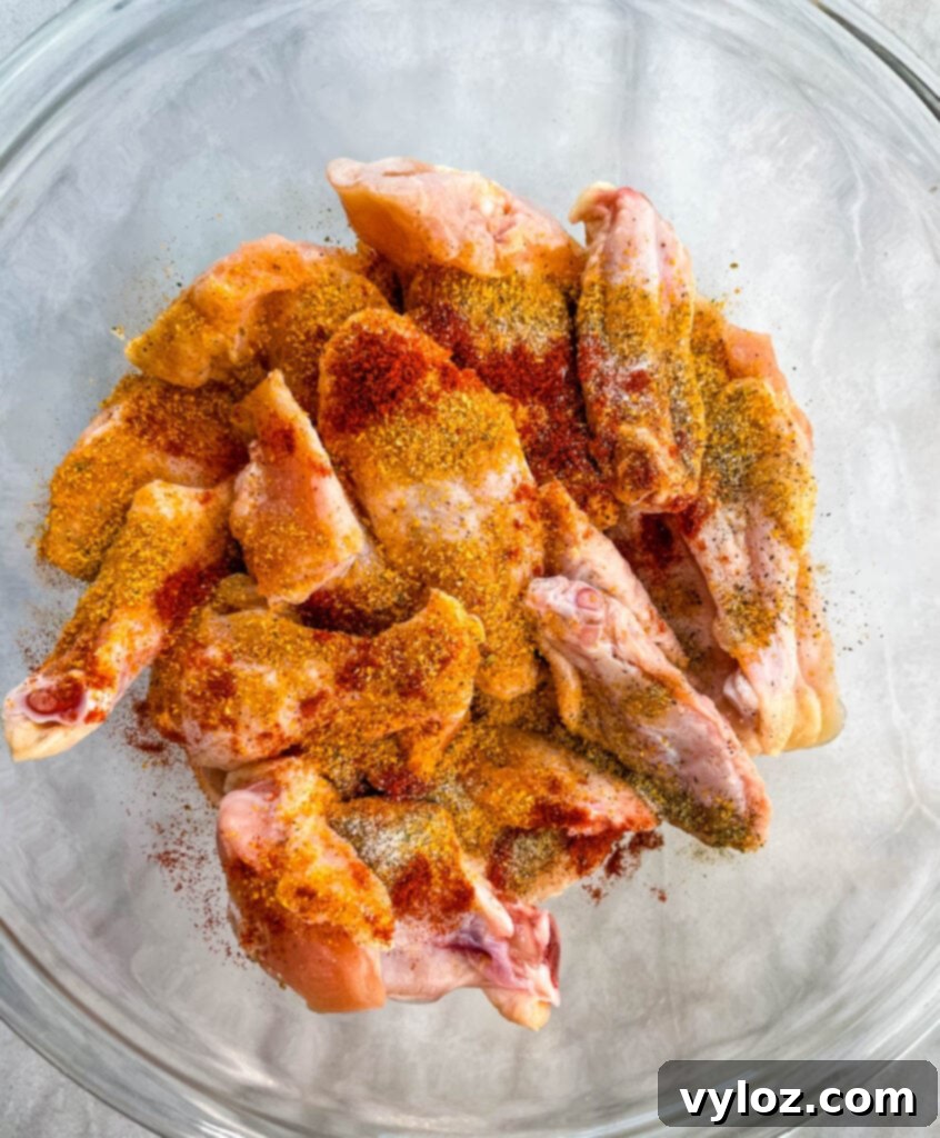 Raw chicken wings generously coated in a dry rub seasoning, ready for cooking, in a glass bowl