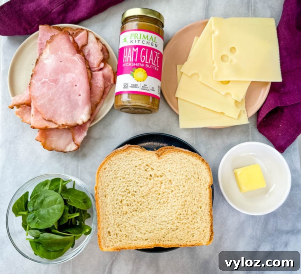 Individual ingredients for a ham and cheese panini laid out: slices of cooked ham, Swiss cheese, two slices of sourdough bread, fresh spinach, and a pat of butter, all on separate plates.