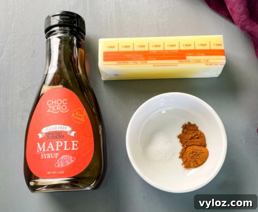A selection of ingredients for maple butter: a stick of butter, a bottle of maple syrup, and a small dish of cinnamon powder arranged on a rustic wooden surface.