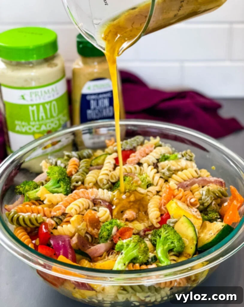 A stream of golden honey dijon mustard drizzling into a vibrant bowl of vegetable pasta salad