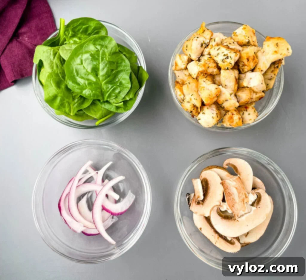 Individual bowls holding fresh spinach, sliced onions, quartered mushrooms, and perfectly cooked, cubed chicken breast, ready to be layered onto pita bread pizzas.