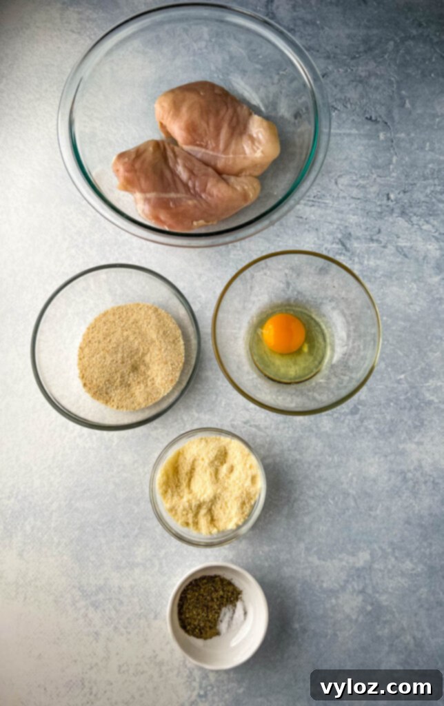 Raw chicken breasts, breadcrumbs, beaten eggs, grated Parmesan cheese, and Italian seasoning laid out in separate bowls, ready for chicken preparation.