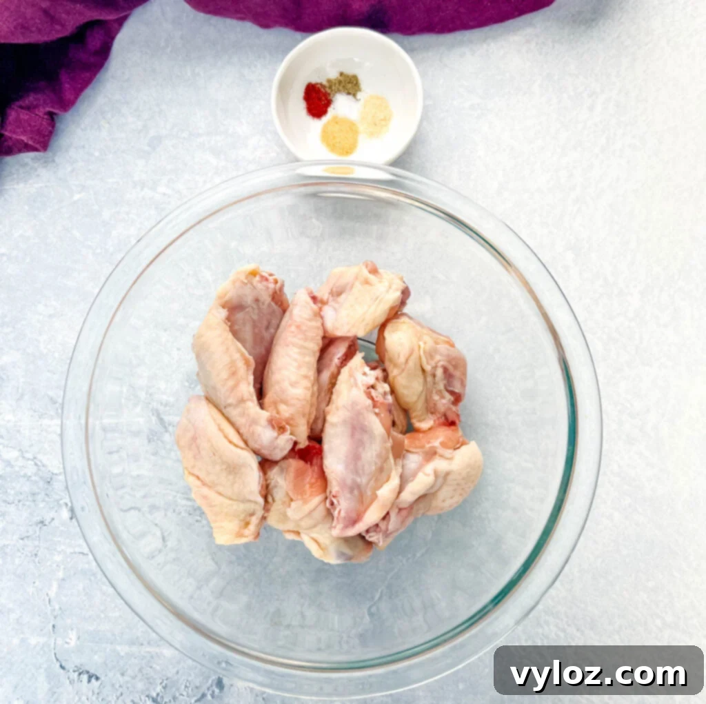 Raw chicken wings in a glass bowl, ready for seasoning, next to a bowl of spices.