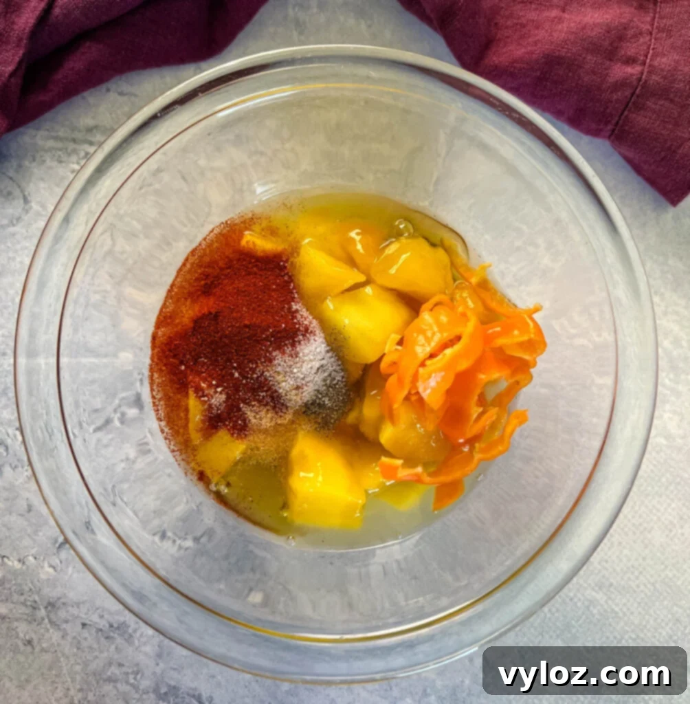 A glass bowl filled with diced mango, whole habanero peppers, honey, and various spices, all ready to be blended into a delicious sauce.