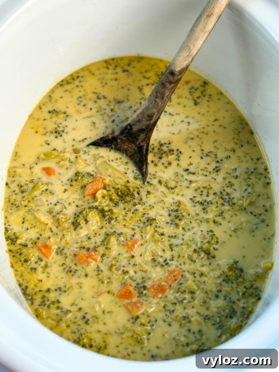 Creamy Slow Cooker Broccoli Cheddar Soup 11 Slow cooker filled with creamy broccoli cheddar soup, showing chunks of broccoli and carrots, with a wooden spoon resting inside.
