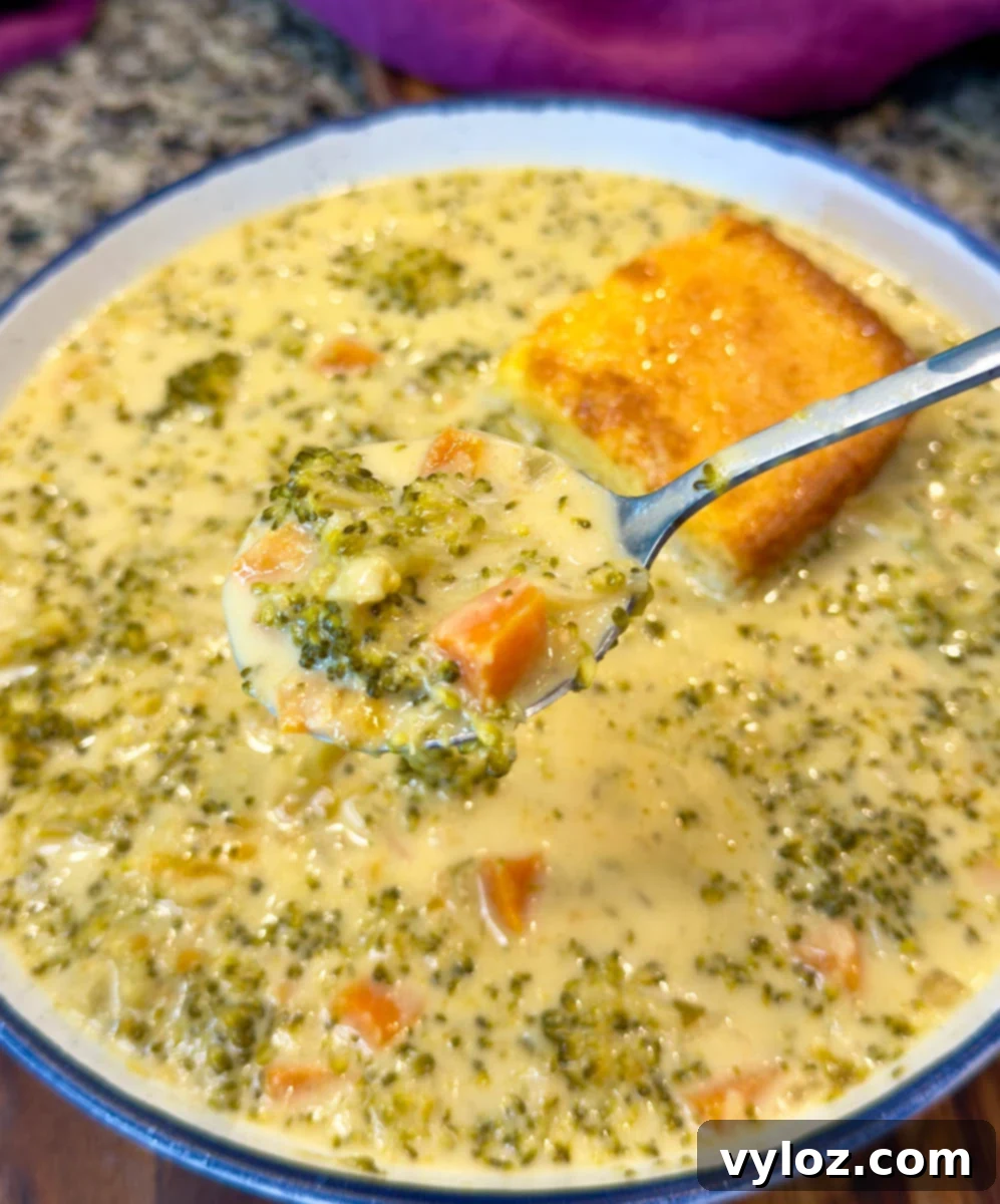 Creamy Slow Cooker Broccoli Cheddar Soup 12 Close-up of a spoonful of broccoli cheddar soup lifted from a bowl, showing tender broccoli and carrots in creamy cheese broth with a square of cornbread in the background.
