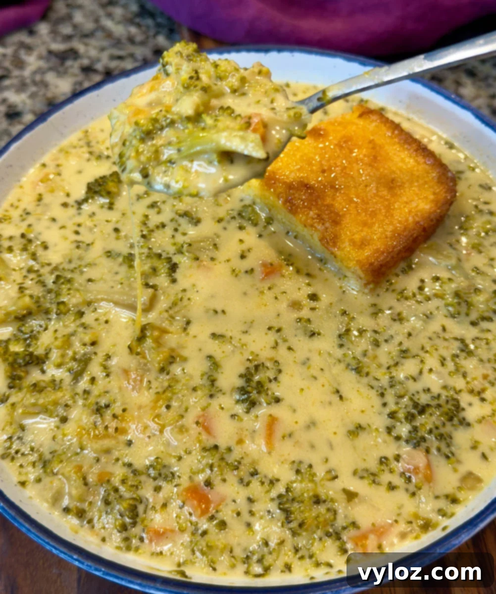 Creamy Slow Cooker Broccoli Cheddar Soup 8 Close-up of a spoonful of cheesy broccoli cheddar soup lifted from a bowl, showing melted cheese strands, tender broccoli, and carrots, with a golden square of cornbread in the soup.