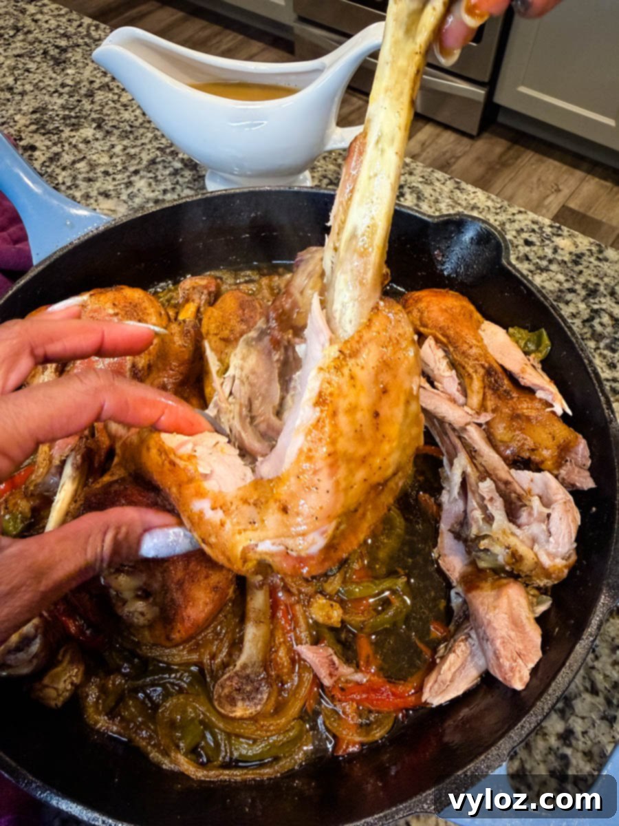 Hands pulling apart a tender baked turkey leg in a cast iron skillet filled with caramelized onions, bell peppers, and rich broth. The meat is visibly falling off the bone, showing how juicy and tender it is, with a gravy boat in the background.