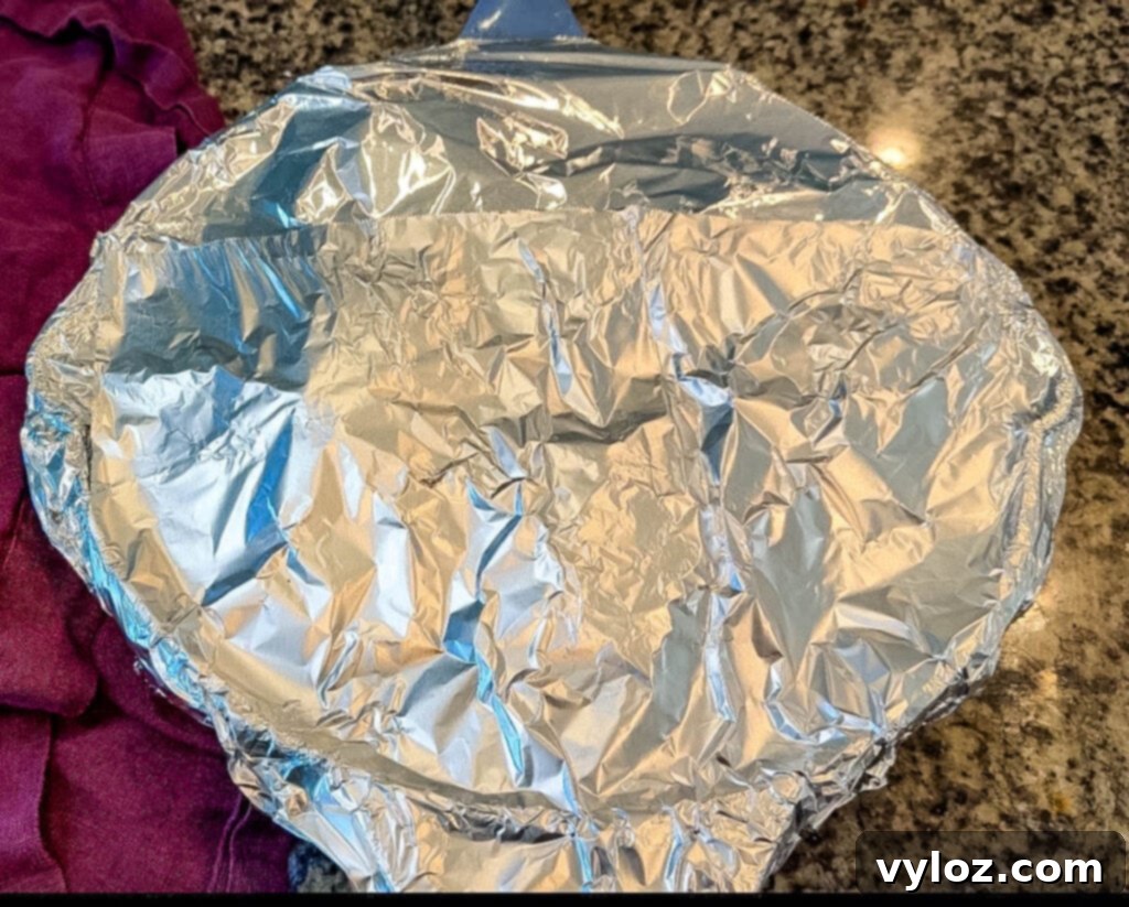 A baking dish covered tightly with aluminum foil, ready for the oven. A purple kitchen towel rests beside it on the countertop.