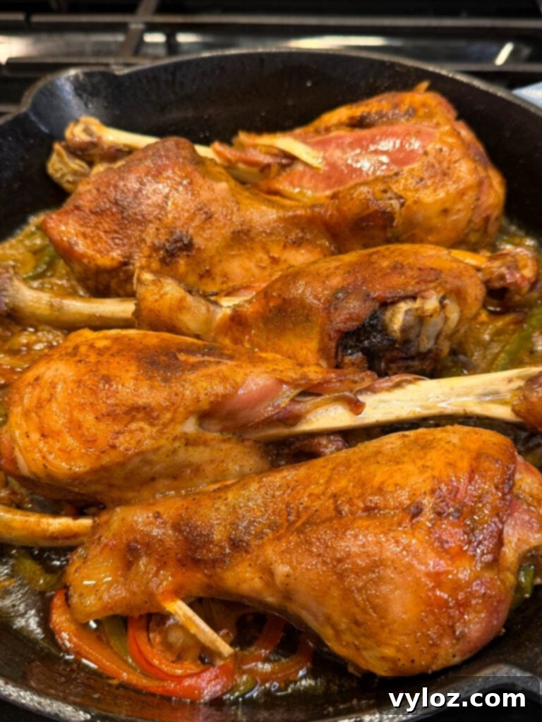 Close-up of baked turkey legs in a cast iron skillet, showing the juicy, seasoned skin and roasted vegetables beneath. The turkey looks tender and fall-off-the-bone, with visible steam and shine from the cooking juices.