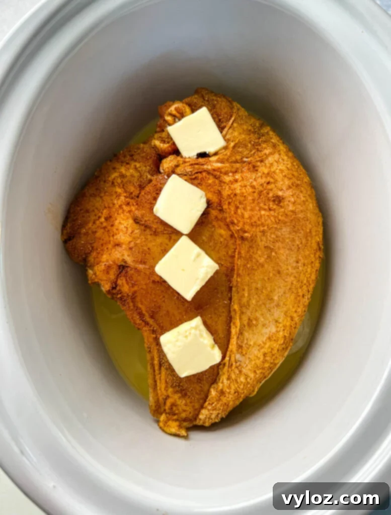 Raw bone-in turkey breast generously seasoned and nestled into a Crockpot slow cooker, topped with pats of butter, ready for cooking